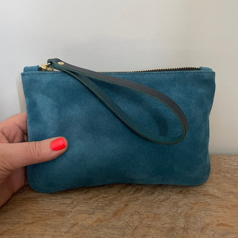Petrol blue suede clutch bag with a strap held by a hand on a wooden surface.